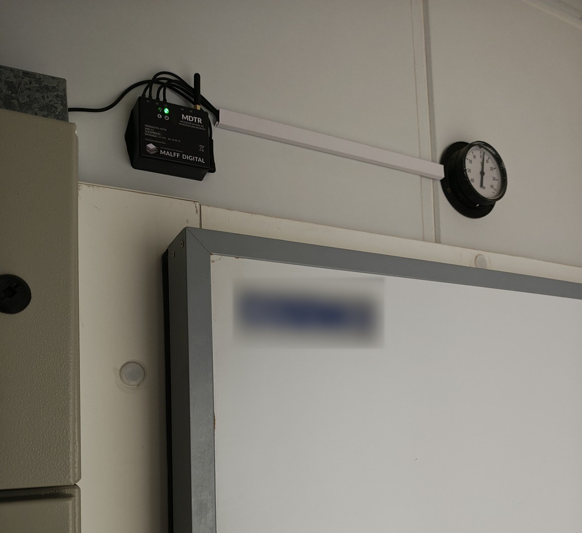 Equipo MDTR instalado en la pared junto a la puerta de una cámara de frío, con termómetro analógico testigo al lado